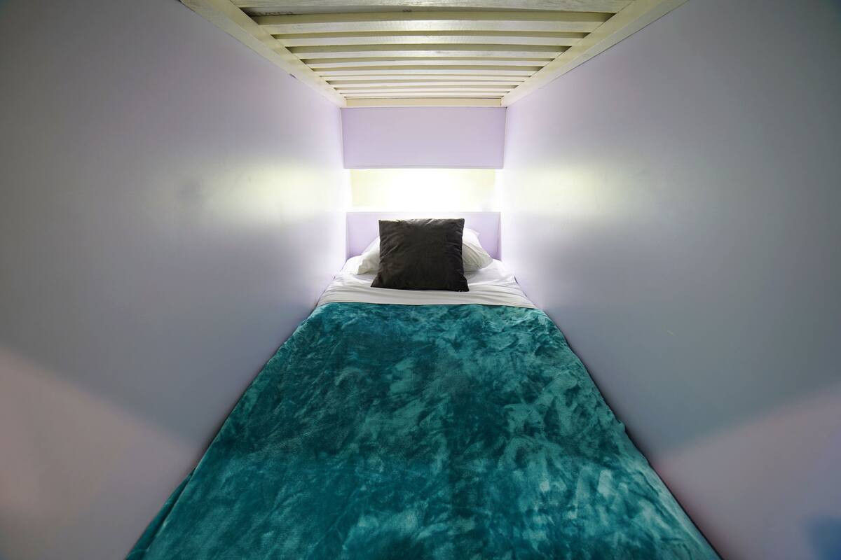 A narrow room with a single bed, a light-colored ceiling, and a small window.