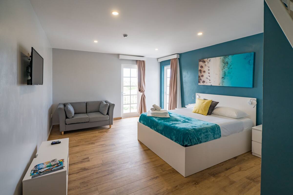 A modern bedroom with a large bed, a grey sofa, and a flat-screen TV mounted on the wall.