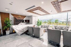 Un vestíbulo moderno de hotel con mostrador de recepción de mármol, zona de estar y grandes ventanales con vistas al exterior.