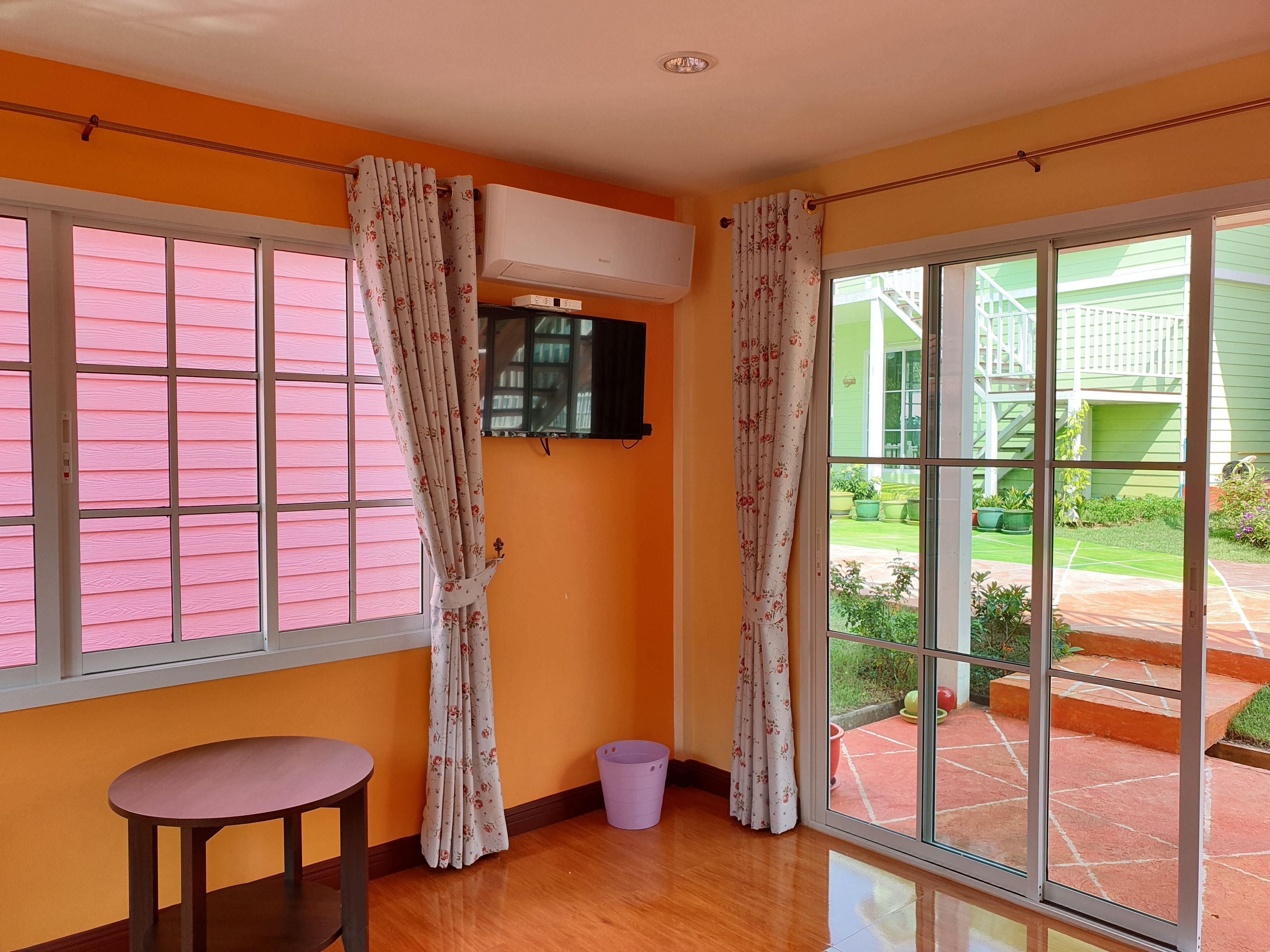 A room with a single table, an air conditioning unit, a window with pink blinds, and a view of a green building outside.