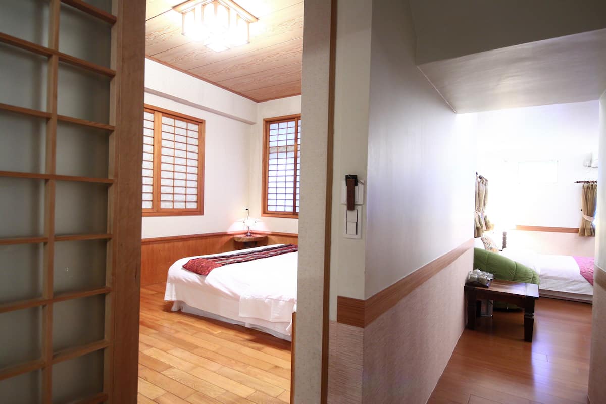 Une pièce de style japonais traditionnel, avec un plancher en bois, un revêtement de sol en tatamis, un lit recouvert d’une courtepointe blanche, une petite table avec une lampe et une porte coulissante.