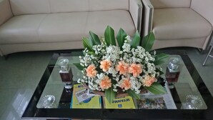 A glass-top table with a floral arrangement, magazines, and a book titled "NYANNA". - Nay Chi Linn Hotel (Pathein)