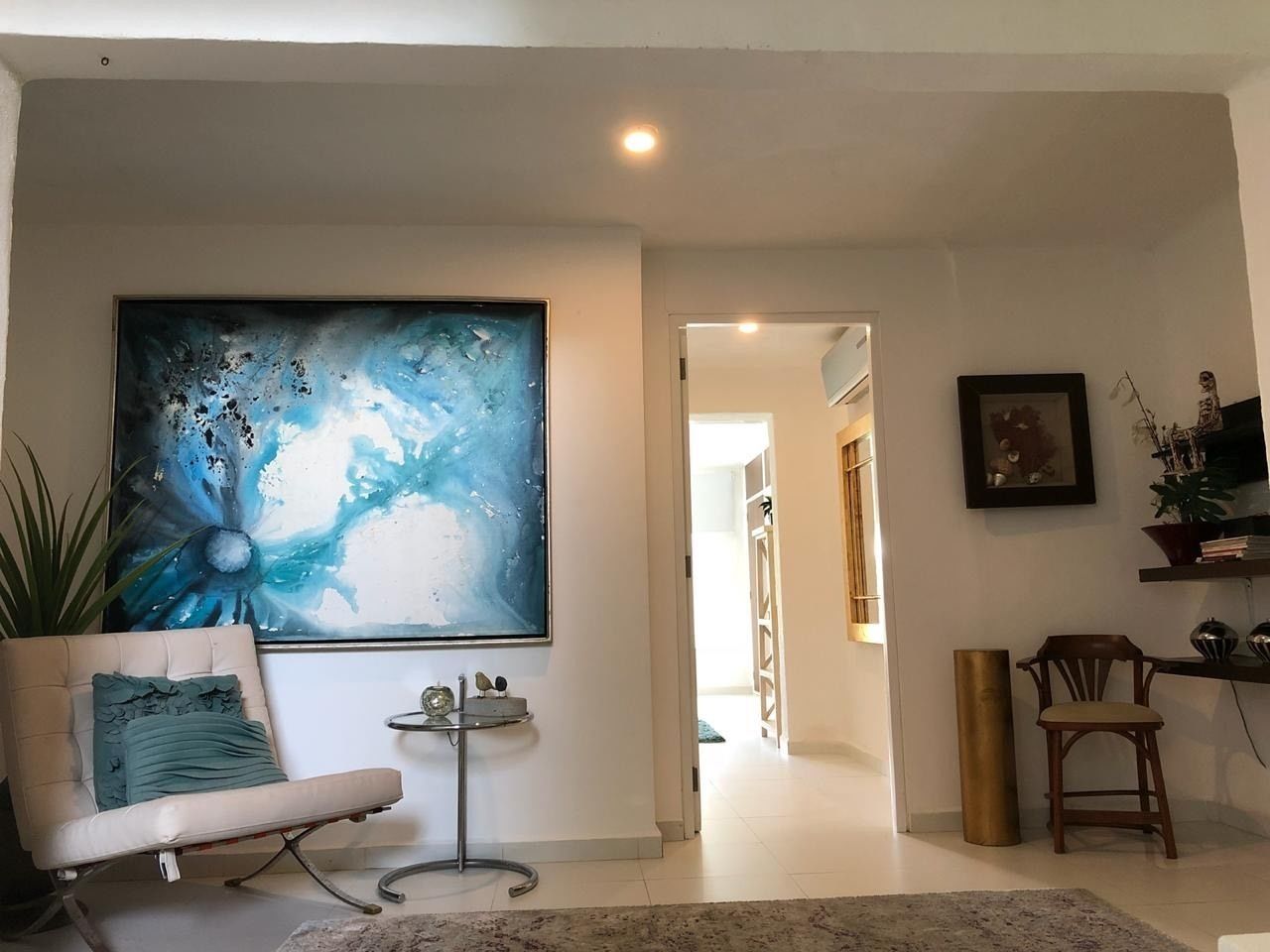 A living room with a large abstract painting, a beige sofa with a teal pillow, a small side table, and a wooden chair.