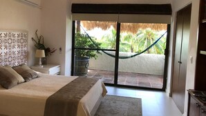 Un dormitorio con cama, mesita de noche, planta en maceta, hamaca y puerta corrediza de vidrio que da a un balcón con vistas a palmeras.