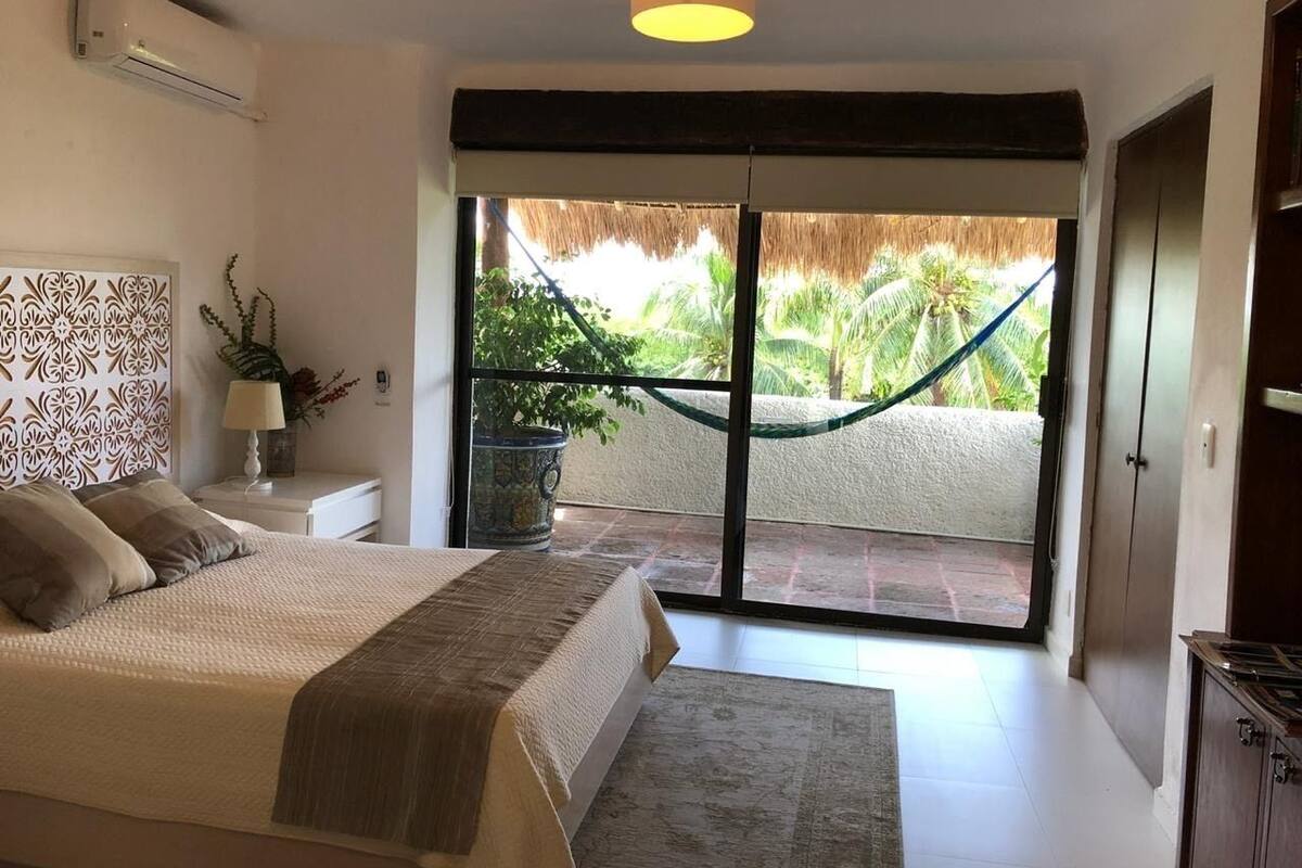 A bedroom with a bed, a nightstand, a potted plant, a hammock, and a sliding glass door leading to a balcony with a view of palm trees.