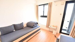 Ein kleines Zimmer mit Schlafsofa, Tisch und einem Vorhangfenster.