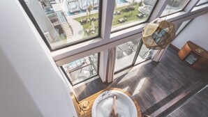 A high-ceilinged room with a large window overlooking a pool area, a wooden floor, and a glass-enclosed jacuzzi.