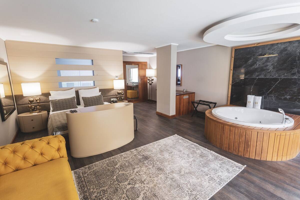 Una habitación de hotel moderna con un sofá curvo, una bañera de madera redonda, una isla central con un lavabo y un espejo grande.