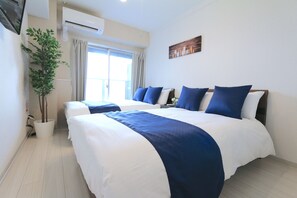 Una habitació d'hotel amb dos llits, una planta en test, un aparell d'aire condicionat i una finestra amb cortines.