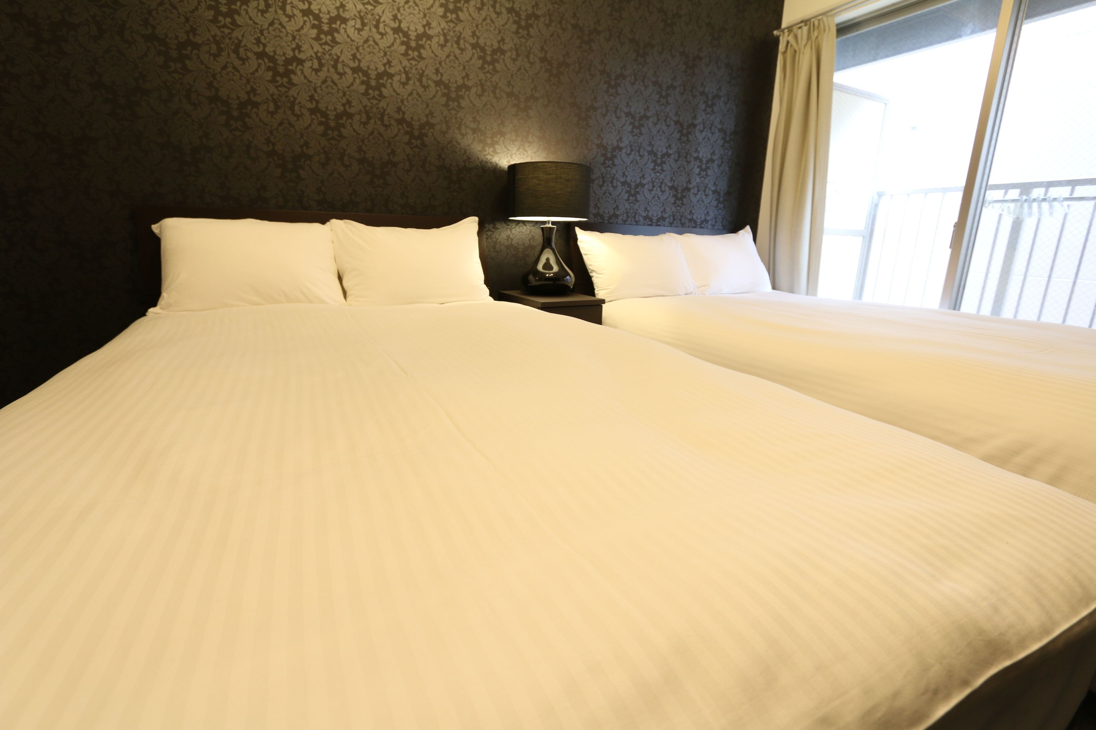 Ein Hotelzimmer mit einem großen Bett, einem Nachttisch mit Lampe und Blick ins Freie durch ein Fenster.