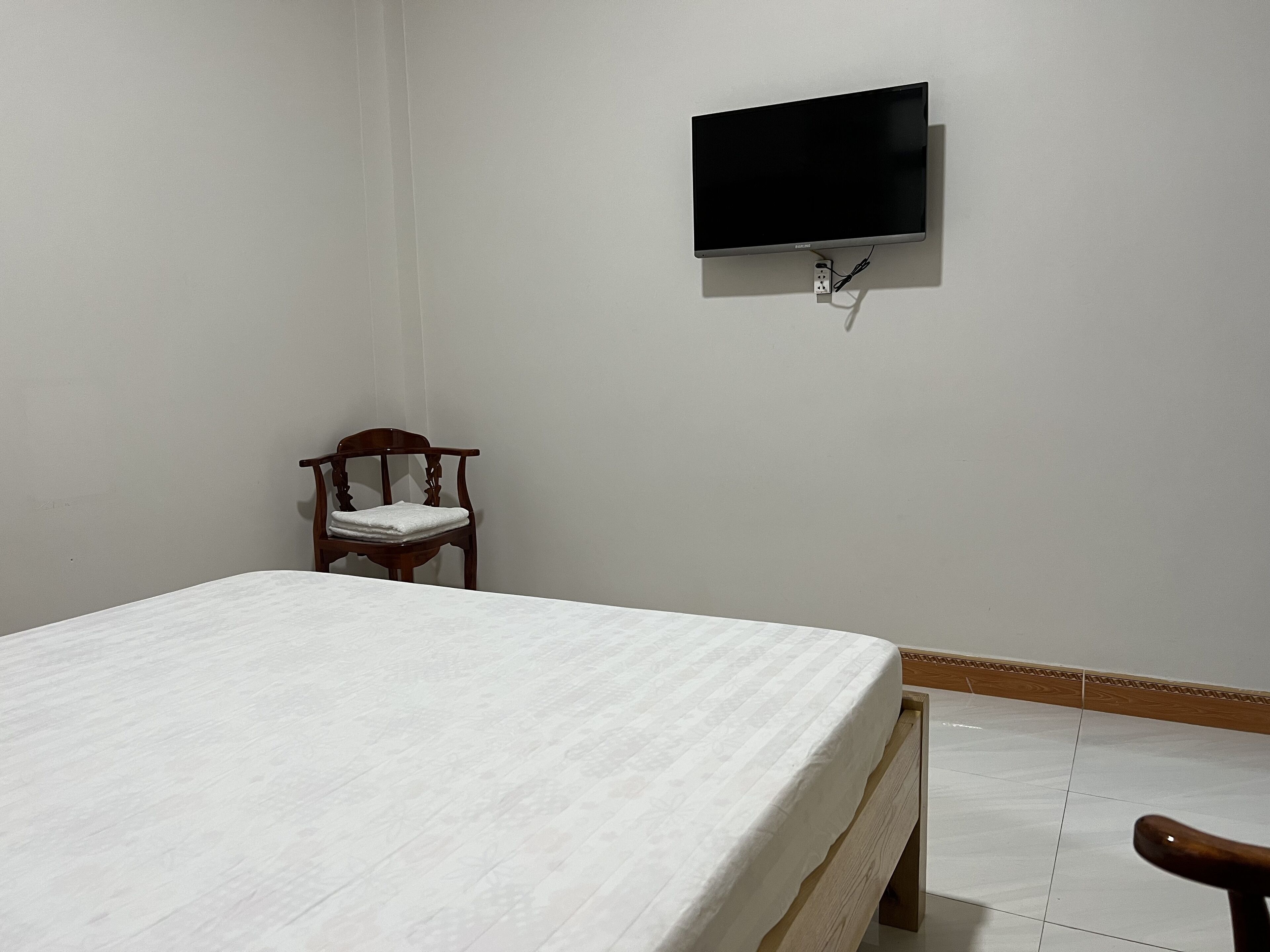 Un dormitorio con una cama, una silla, un televisor montado en la pared y un enchufe.