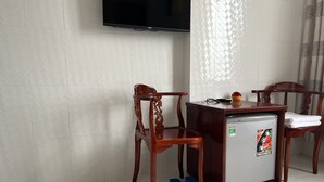 Ein kleines Zimmer mit Holztisch, zwei Stühlen, einem an der Wand montierten Fernseher und einem Kühlschrank mit Aufkleber.