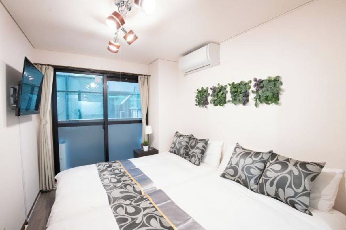 A hotel room with a large bed, a TV on the wall, a window with curtains, and a wall-mounted air conditioner.
