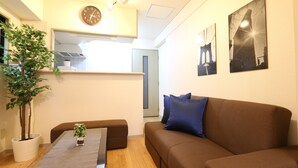 A living room with a brown sofa, a small table with a plant, a clock on the wall, and two framed pictures. - Elizabeth Garden II (Osaka)