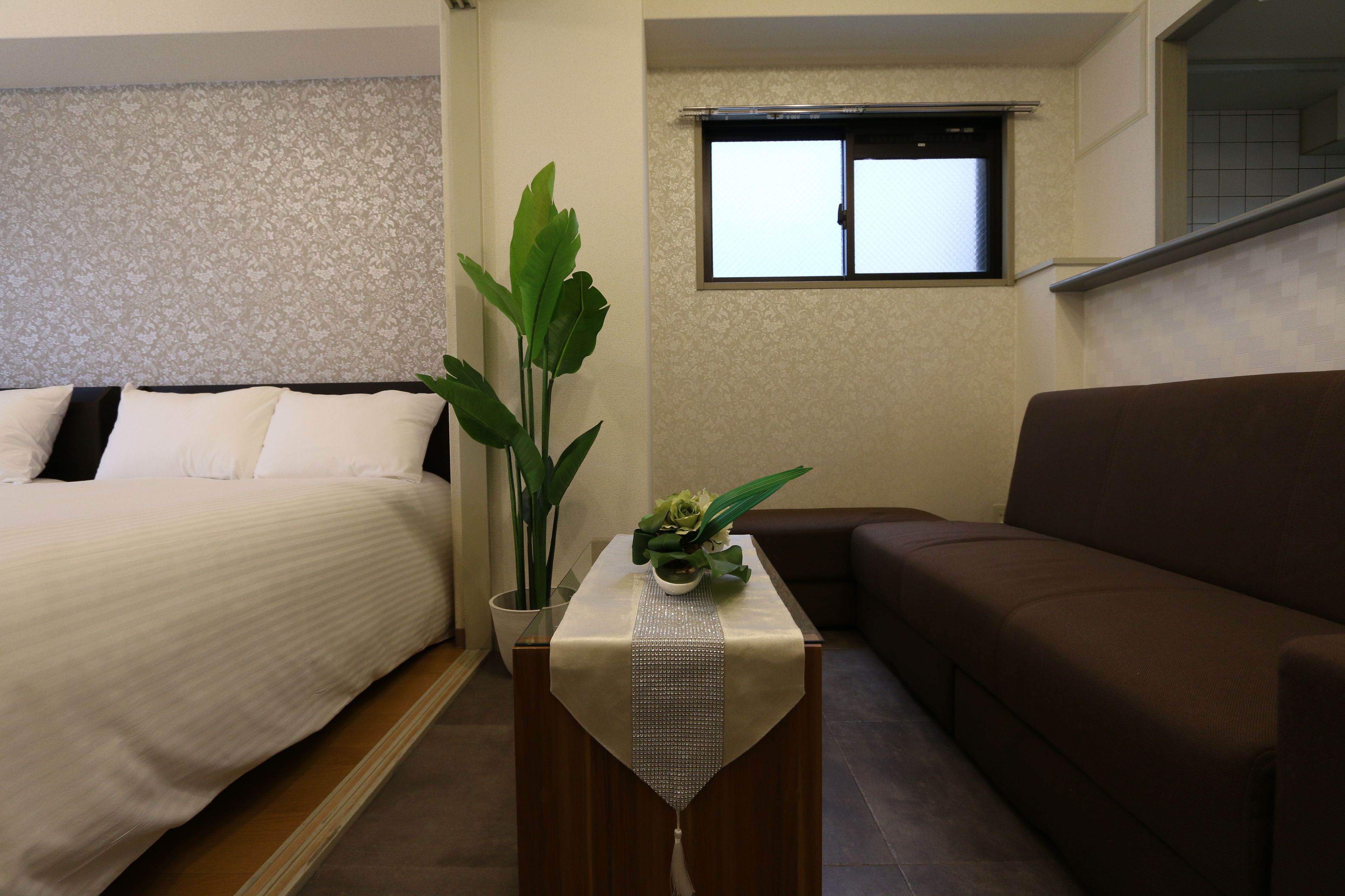 Een slaapkamer met een bed, een bruine bank, een tafeltje met een plant en een raam.