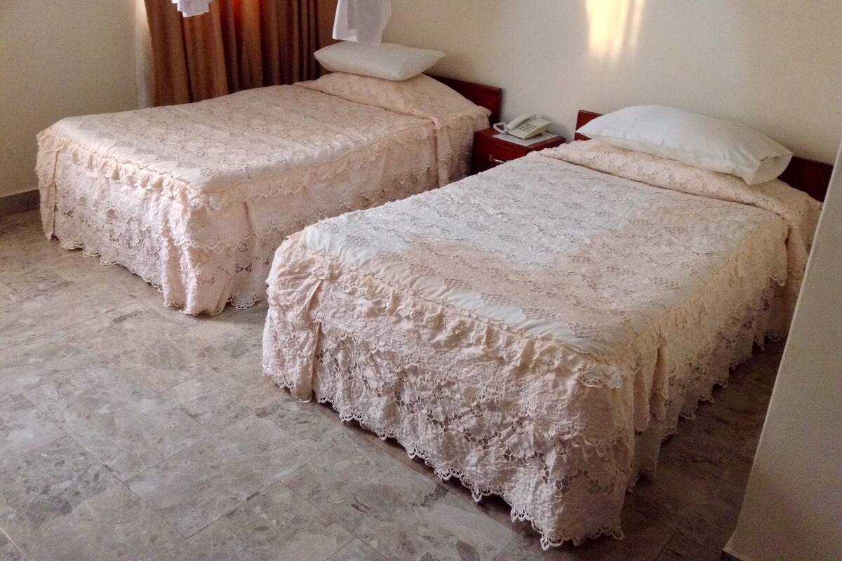 Two single beds with lace covers, a bedside phone, and a curtain in a hotel room.