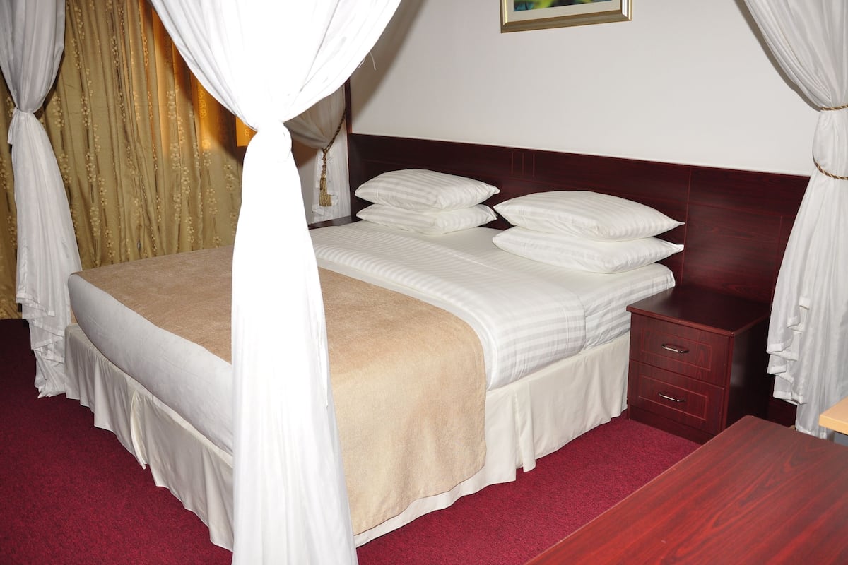 A neatly made bed with a canopy, a wooden nightstand with drawers, and a red carpeted floor.