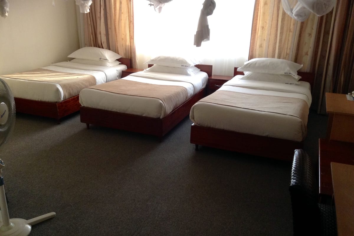 A hotel room with four single beds arranged in a row, a standing fan, and a wooden nightstand with a lamp.