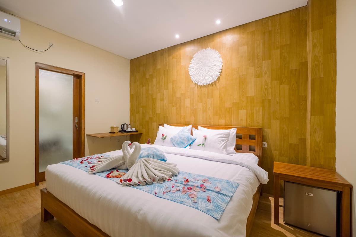 Quarto de hotel com cabeceira de madeira, cama com roupa de cama branca, ar condicionado fixado na parede, pequena mesa de cabeceira com abajur e piso de madeira.