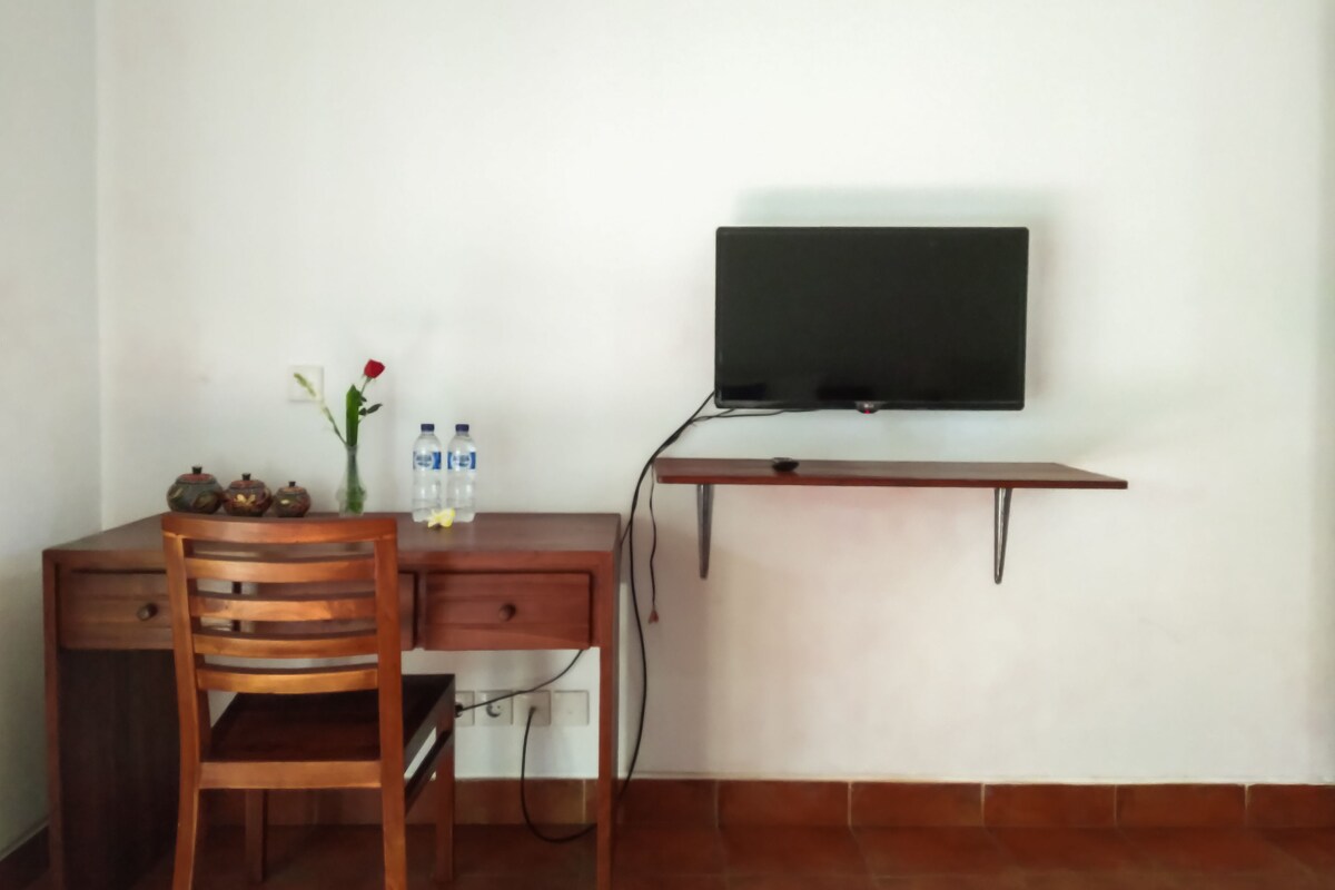 Une petite pièce simple, avec un bureau en bois, une chaise et un téléviseur fixé au mur. Sur le bureau, on trouve deux bouteilles d’eau et un vase contenant une seule rose rouge.