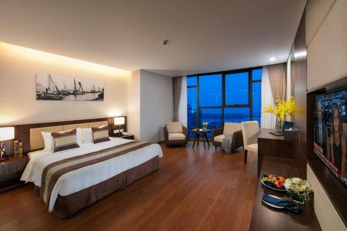 A hotel room with a large bed, two armchairs, a small table with a vase, and a large window with a city view.