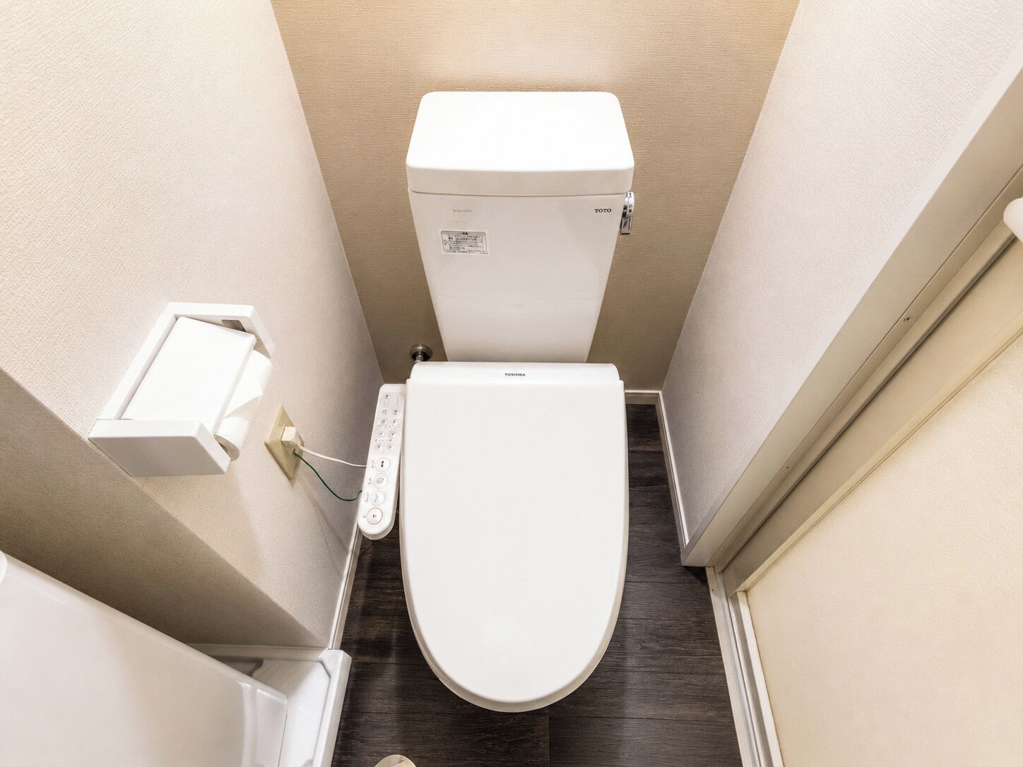 A compact bathroom with a white toilet, a wall-mounted electrical outlet, and a small trash bin.