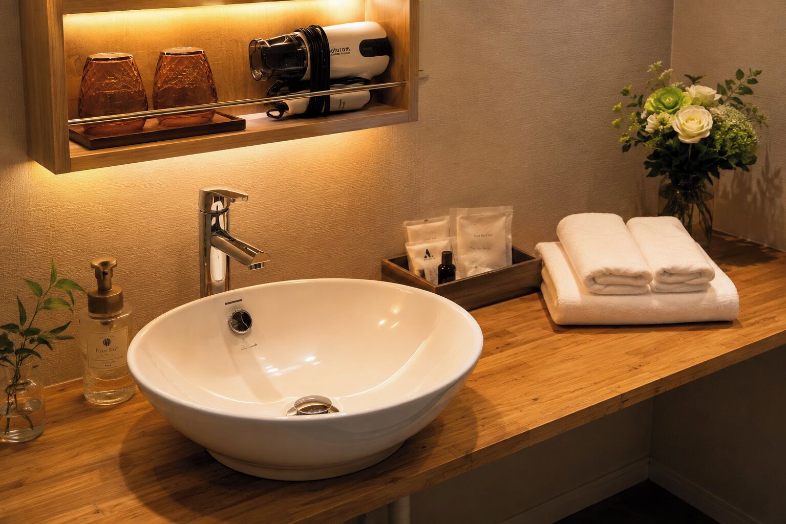Un baño de hotel con un lavamanos grande de color blanco, un espejo encima, un secador de pelo en un estante, un florero con flores y toallas dobladas cuidadosamente.