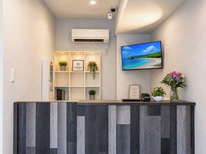 Un mostrador de recepción con un televisor de pantalla plana que muestra una escena de playa, una planta pequeña y un cuadro en la pared.
