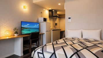 Une chambre d’hôtel compacte comprenant un lit, un bureau avec un ordinateur, une petite kitchenette et un téléviseur fixé au mur.