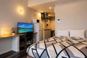 Habitación de hotel pequeña con cama, escritorio con computadora, kitchenette y televisor montado en la pared.