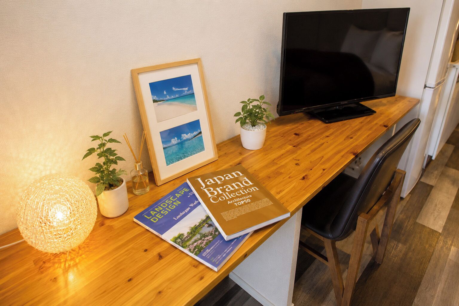 Un escritorio de madera con un cuadro enmarcado, un libro titulado 'Japan Brand Collection Architecture TOP50', un televisor y una planta en maceta.