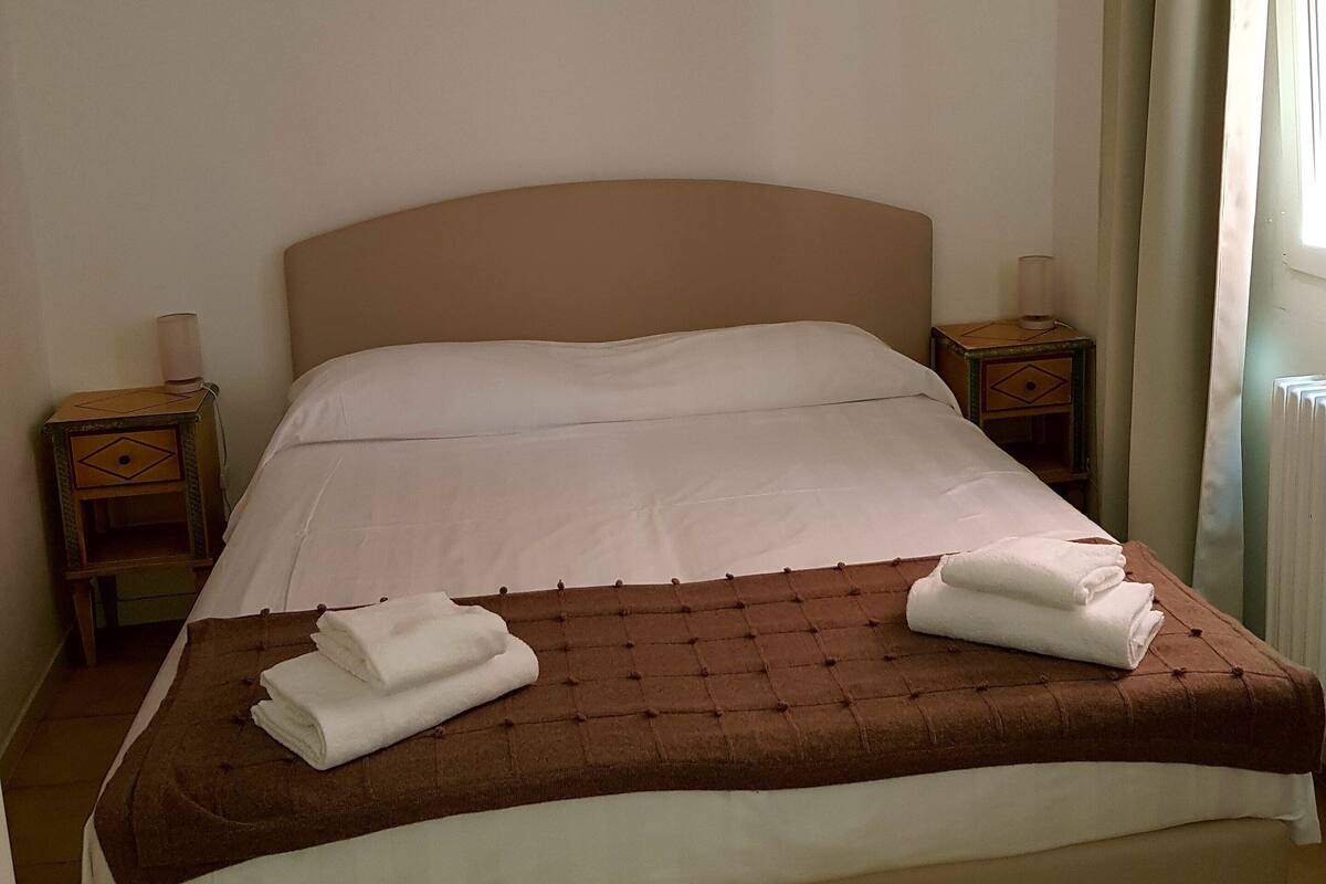 A neatly made bed with a brown headboard, white linens, and two folded towels on the footboard. There are two bedside tables with lamps, and a window with curtains on the right.