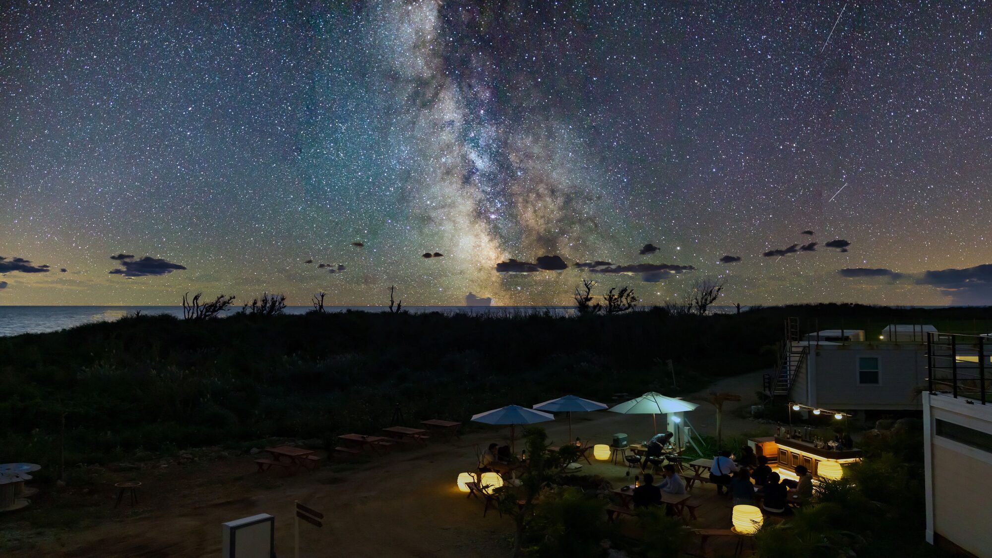 A starry night sky with the Milky Way visible, a beach setting with tents and tables, and a distant view of the ocean.