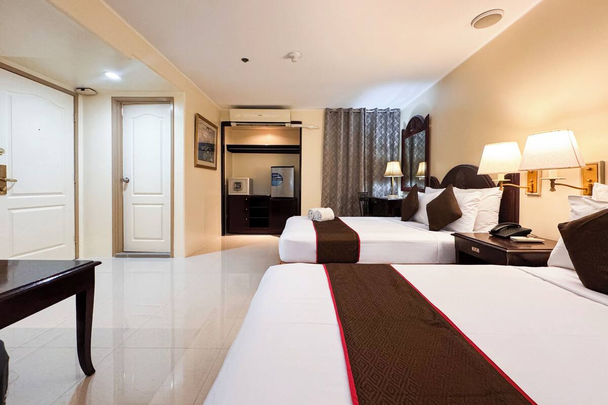 Une chambre d’hôtel avec deux lits, un bureau, une chaise, une petite table et une salle de bain visible à travers une porte ouverte.