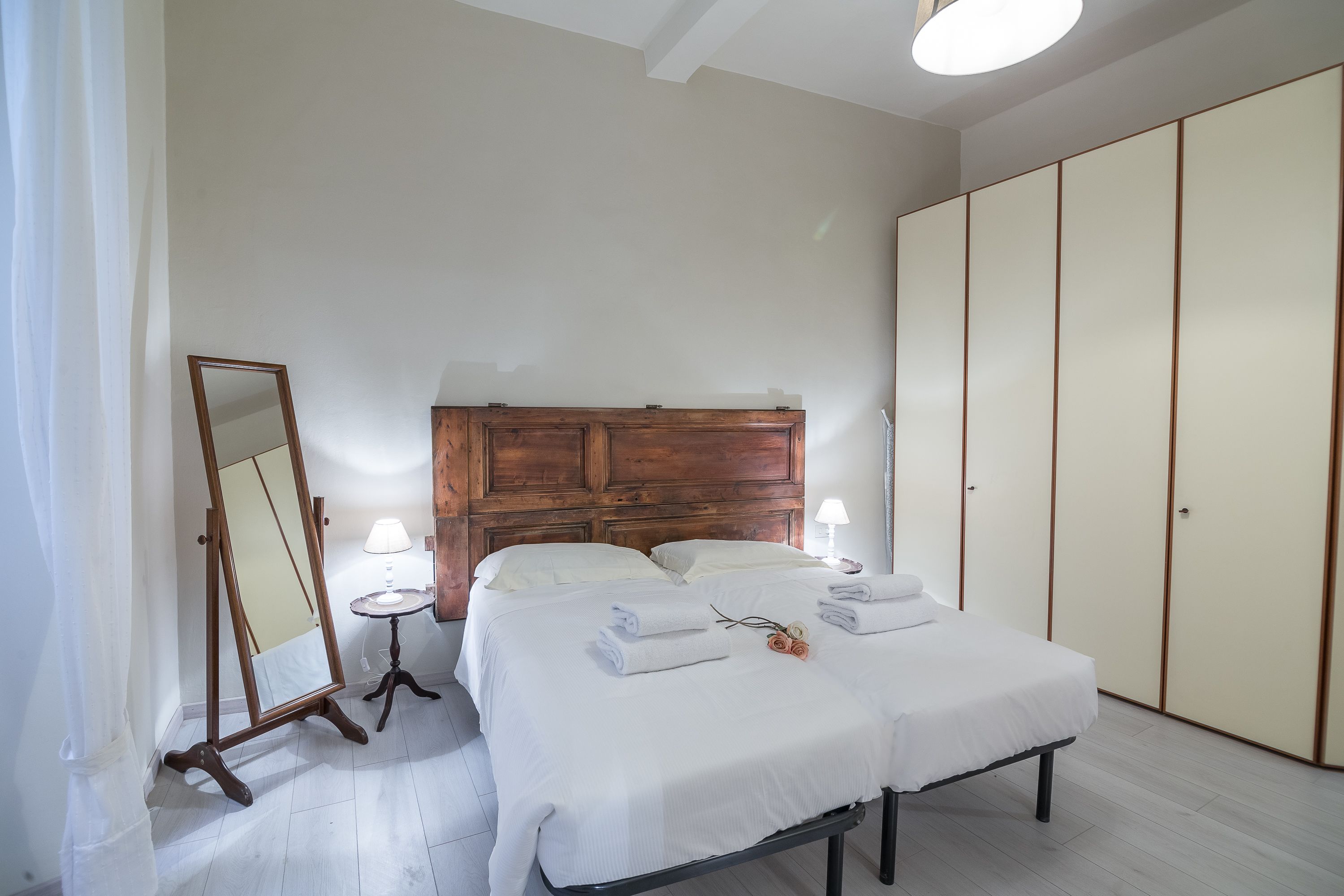 A bedroom with a wooden headboard, two beds, a bedside table with a lamp, and a wardrobe.