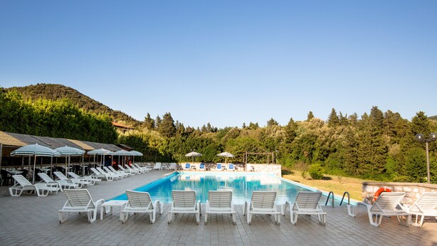 A pool area with white lounge chairs and umbrellas, surrounded by a stone wall and a forested hillside. - Villa Rinascimento Dependance (Lucca)