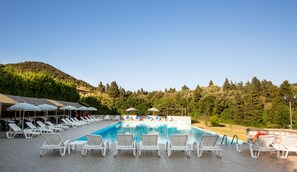 A pool area with white lounge chairs and umbrellas, surrounded by a stone wall and a forested hillside.
