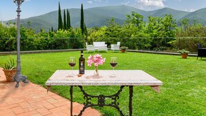 A table with a bottle and glasses, set against a backdrop of a grassy garden and mountains. - Villa Rinascimento Dependance (Lucca)