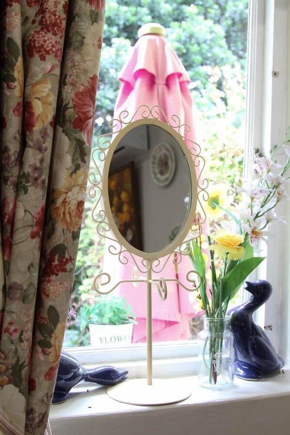 A window with a floral curtain, a decorative mirror, a vase with flowers, and a pink umbrella outside.