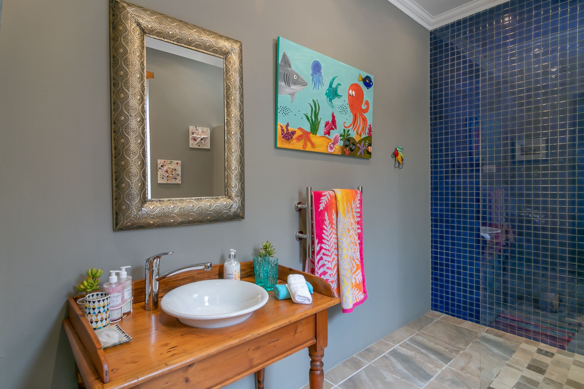 A bathroom with a wooden vanity, a white sink, a gold-framed mirror, and a colorful painting of sea creatures.