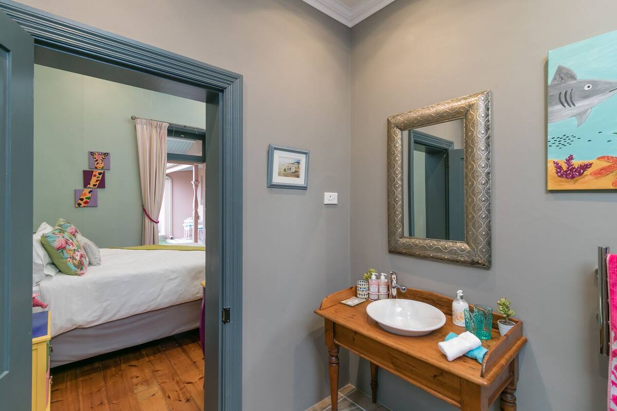 Een slaapkamer met een bed, een houten dressoir met een wastafel en een spiegel. Er is een deur naar een andere kamer.