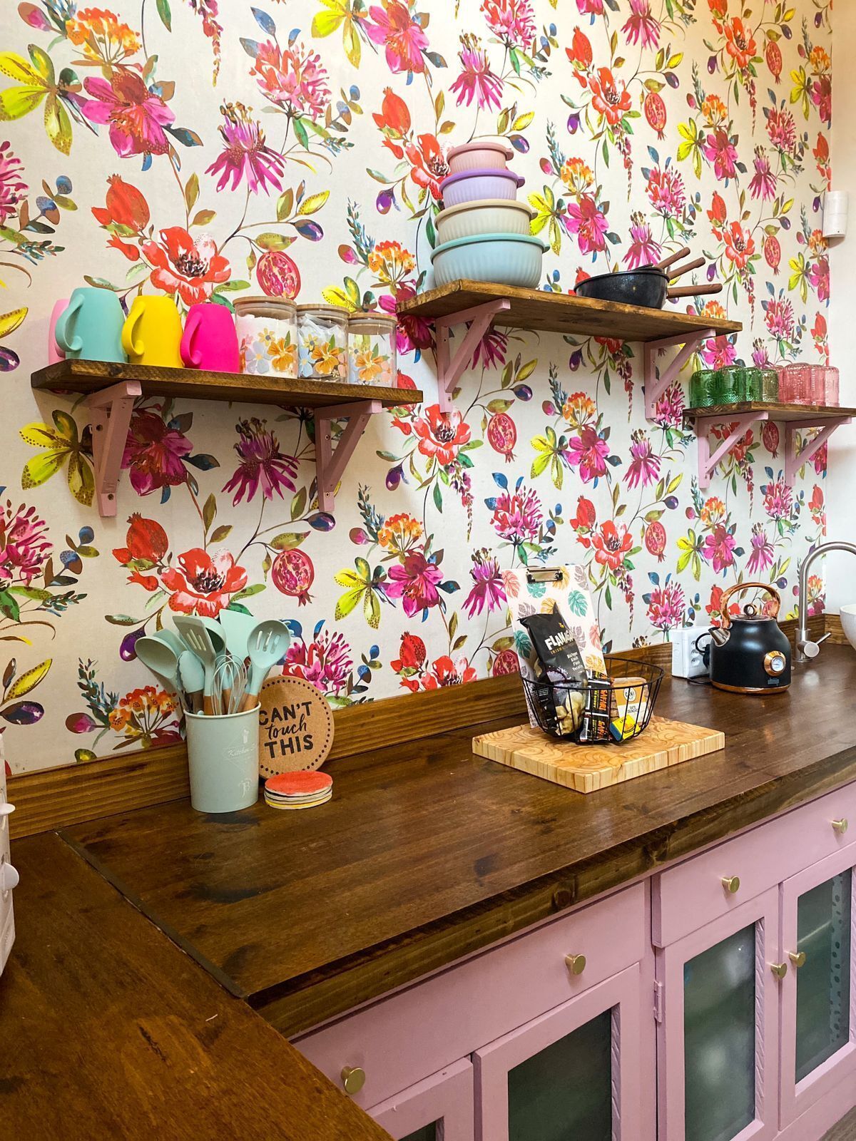 A kitchen with wooden cabinets, a wooden countertop, and a floral wallpaper. There are various kitchen utensils and a kettle on the countertop.