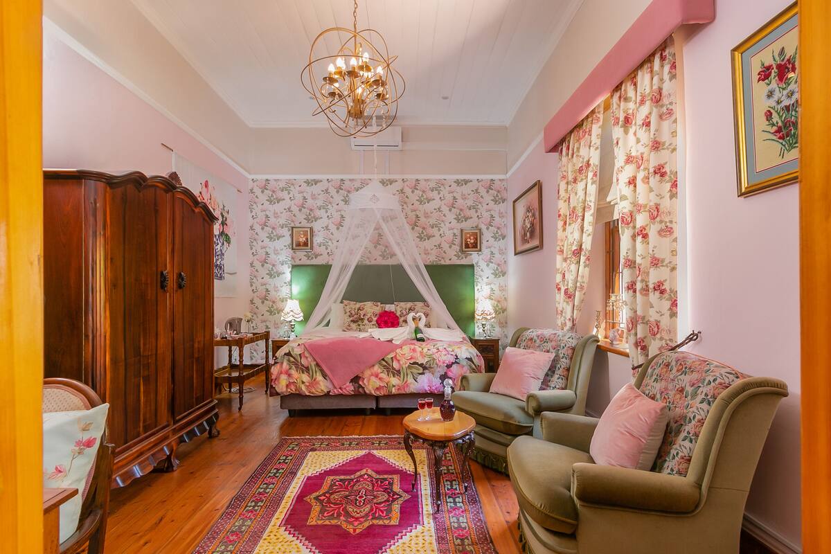 Een slaapkamer met hemelbed, bloemenbehang, een houten garderobe, een groene fauteuil en een vloerkleed met dessin.