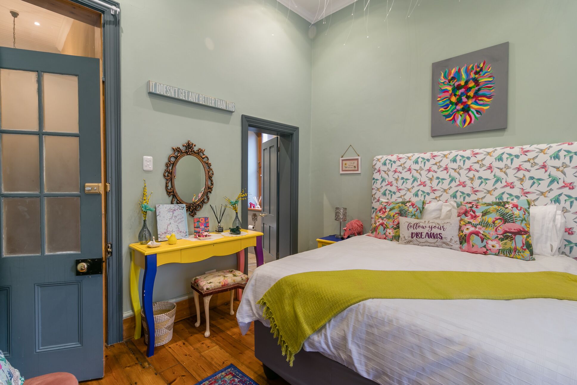 Een slaapkamer met een bed, een dressoir, een spiegel en een muur met kleurrijke kunstwerken.