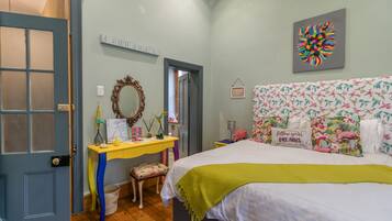 Een slaapkamer met een bed, een dressoir, een spiegel en een muur met kleurrijke kunstwerken.