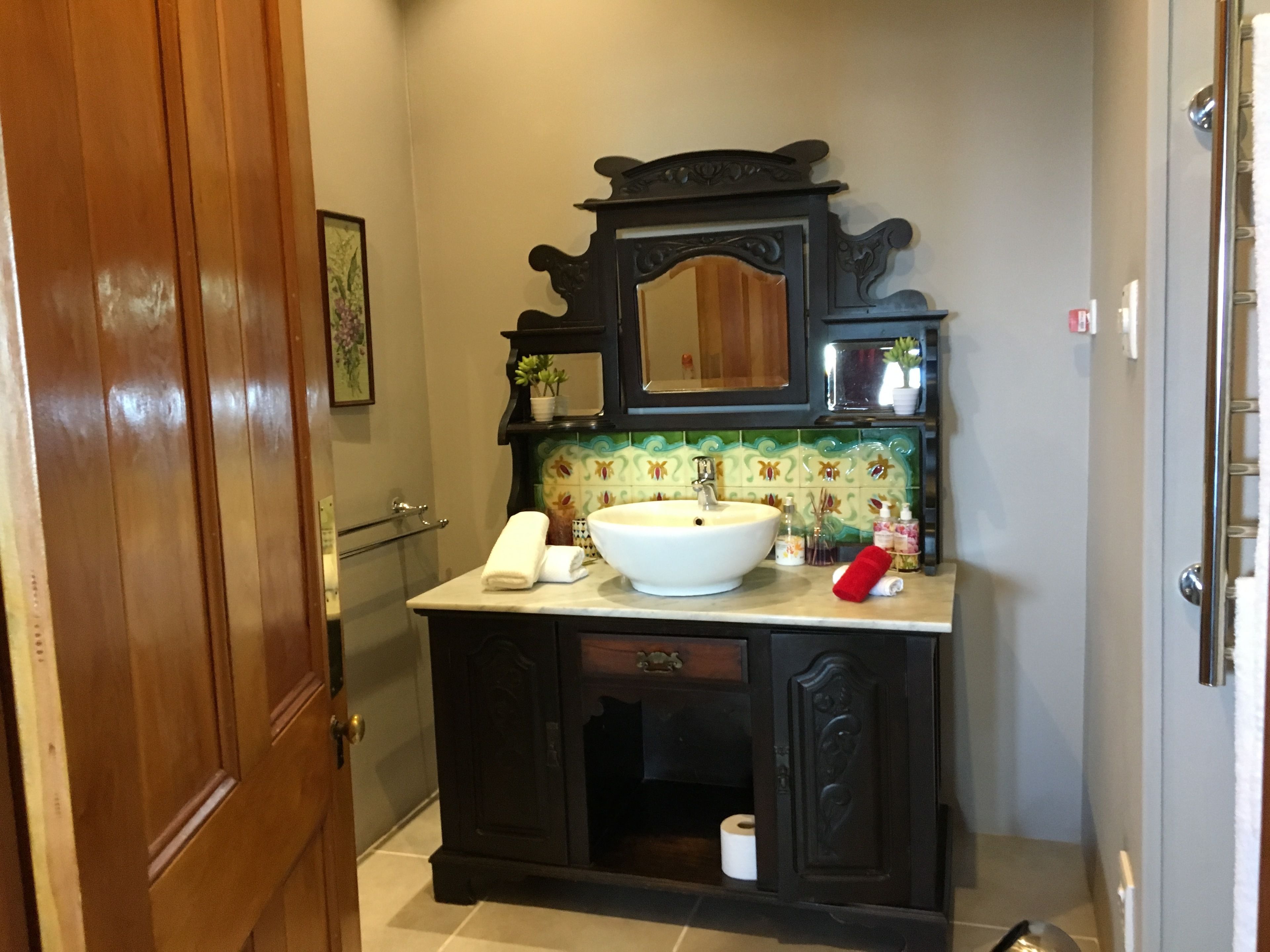 A bathroom with a wooden vanity, a large white sink, and a mirror above it. There is a towel rack and a toilet paper holder on the vanity.