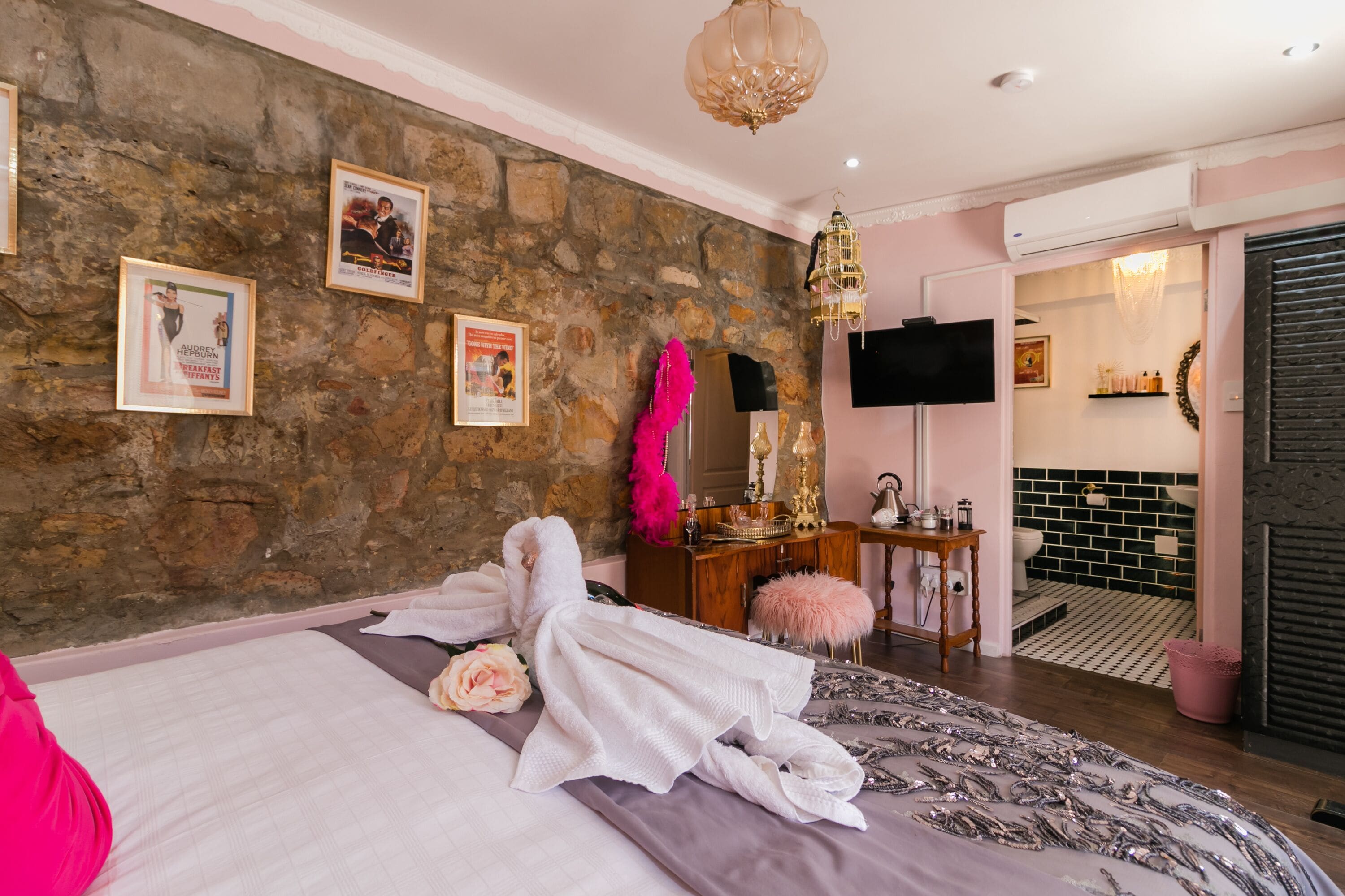 A bedroom with a stone wall, a bed with white and pink linens, a vanity with a mirror, and a television on the wall.