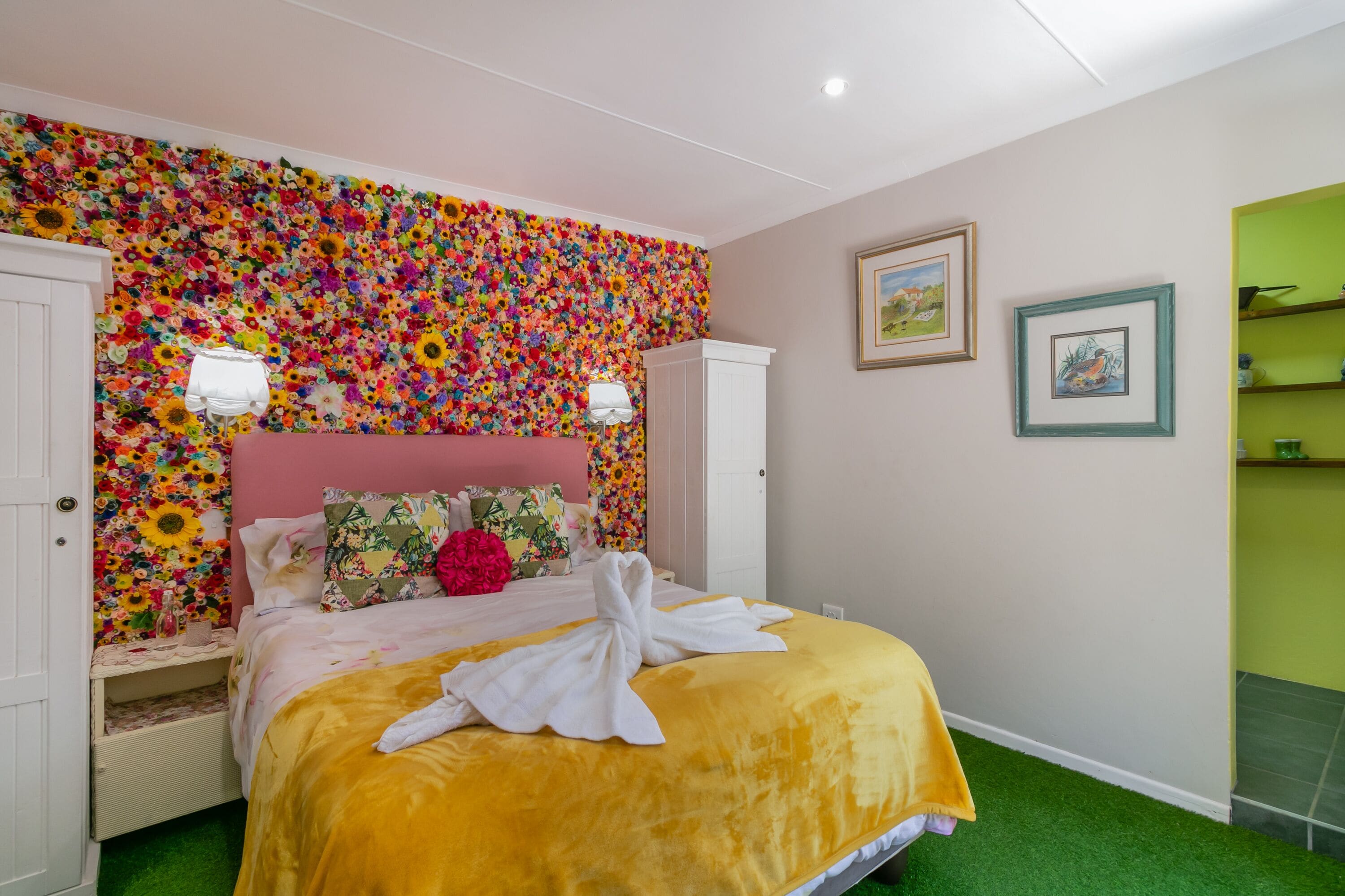 Habitación de hotel con un mural floral en la pared, una cama con cubrecama amarillo y una toalla blanca sobre la cama.