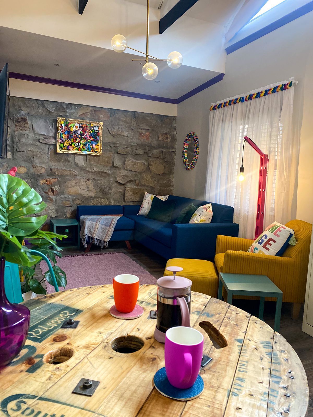 Una sala de estar acogedora con un sofá azul, un sillón amarillo, una mesa de centro de madera con tazas y una pared de piedra con una obra de arte colorida.