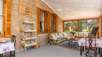 A cozy room with a stone wall, wooden shutters, a sofa, a table, and a window with a view of greenery.